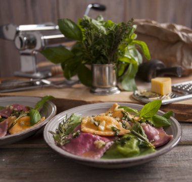 Ahşap bir masada ravioli makarna ile gri bir tabak vardır. Arka planda haddeleme hamur ve baharatlı otlar bir buket için bir makinedir. Sırada bir parça peynirli bir rende var..