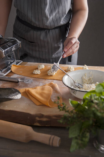 A girl in a gray apron at a wooden table lays out the stuffing on the dough. In the foreground lies a rolling pin and a bouquet of spicy herbs.