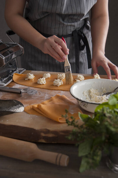 A girl in a gray apron at a wooden table moistens rolled out dough with a brush. In the foreground lies a rolling pin and a bouquet of spicy herbs.