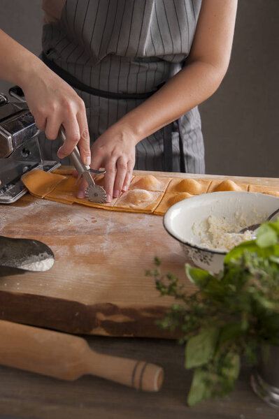 A girl in a gray apron at a wooden table cuts ravioli paste out of dough. In the foreground lies a rolling pin and a bouquet of spicy herbs.