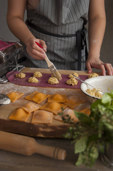 A girl in a gray apron at a wooden table wets rolled out dough with a brush. In the foreground lies a rolling pin, a bouquet of herbs and cooked ravioli pasta.