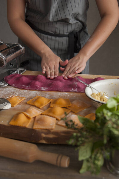 A girl in a gray apron at a wooden table makes ravioli paste from dough. In the foreground lies a rolling pin, a bouquet of herbs and cooked pasta.