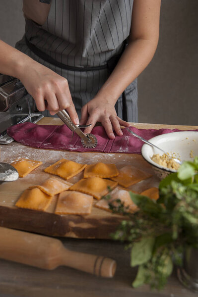 A girl in a gray apron at a wooden table cuts ravioli paste out of dough. In the foreground lies a rolling pin, a bouquet of herbs and cooked pasta.