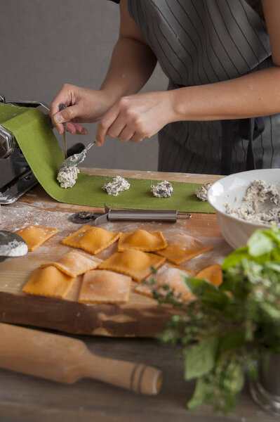 A girl in a gray apron at a wooden table lays out two spoons of filling on a green dough. In the foreground lies a rolling pin, a bouquet of herbs and cooked pasta.