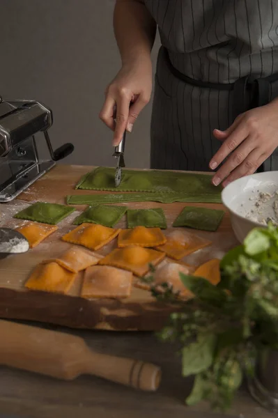 Tahta bir masada gri önlük içinde bir kız yeşil hamurdan ravioli makarna keser. Ön planda bir oklava, otlar ve pişmiş makarna bir buket yatıyor.