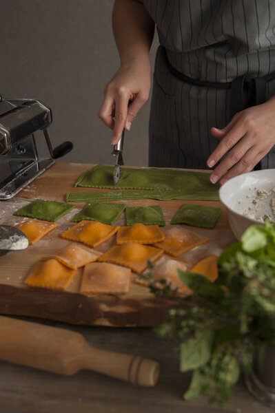 A girl in a gray apron at a wooden table cuts ravioli pasta out of green dough. In the foreground lies a rolling pin, a bouquet of herbs and cooked pasta.