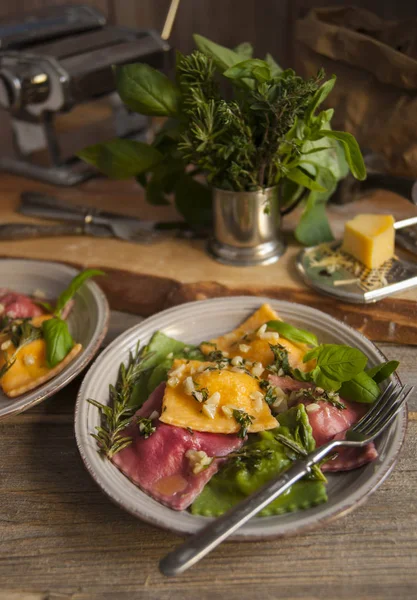 Op een houten tafel is er een grijze plaat met ravioli pasta en een vork. Op de achtergrond is een boeket van kruidige kruiden. Vervolgens is een rasp met een stukje kaas. — Stockfoto