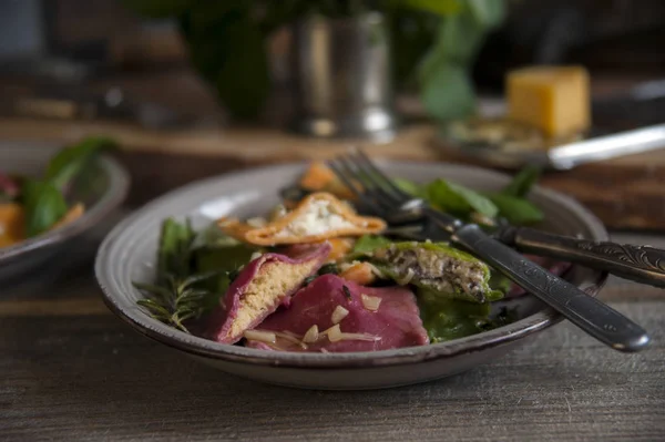 Su un tavolo di legno c'è un piatto grigio con ravioli e posate. Sullo sfondo c'è una grattugia con un pezzo di formaggio . — Foto Stock