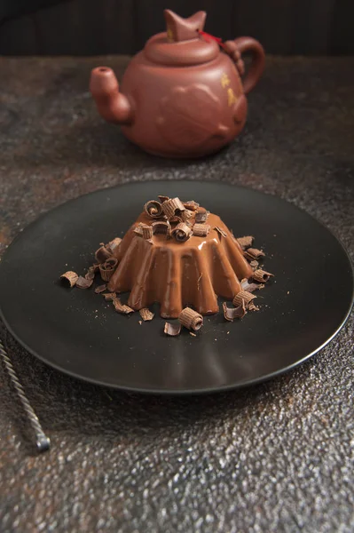 茶色のテーブルの上に黒いプレートがあり、チョコレートパナコタにチョコレートチップを振りかけます。近くにセラミックティーポットがあります. — ストック写真