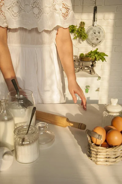 The girl in a white lace dress at the table holding a rolling pin. On the table are dishes and ingredients for making dough. Behind on a brick wall colander.