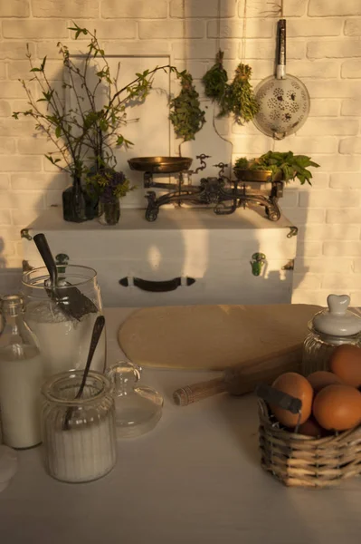 Provence style kitchen is lit by the evening sun. On the table, the dough and dishes with ingredients. In the background, a white brick wall has a chest with weights and herbs.