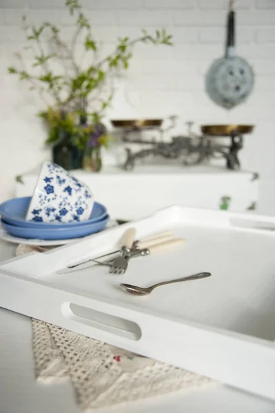 In the Provence-style kitchen on the table is a tray with dishes and cutlery. There is a chest by the white brick wall. On it are scales and vases of flowers. Behind the wall is a colander.