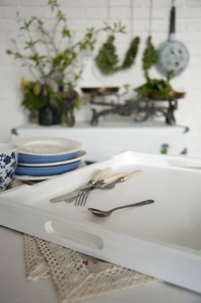 In the Provence-style kitchen on the table is a tray with dishes and cutlery. There is a chest by the white brick wall. There are scales and vases with flowers and greens on it.