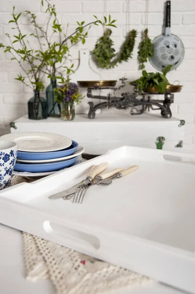 In the kitchen in the style of Provence a white brick wall stands a chest. There are scales and vases with flowers and greens on it. In the front on the table is a tray with dishes and cutlery.