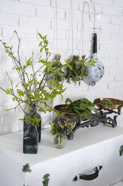 There are vintage scales and vases with bouquets and branches by a white brick wall on a suitcase. Behind them hang bouquets of herbs and colander.