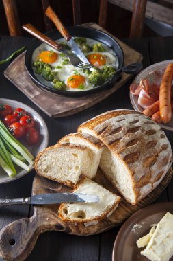 Kahvaltının servis edildiği siyah ahşap masanın üst görünümü: kızarmış yumurta, sosis, sebze, ev yapımı ekmek ve tereyağı.