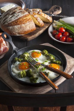 Kahvaltı siyah ahşap bir masanın üzerine serilir: tavada brokoli ile kızarmış yumurta, sebze, ev yapımı ekmek ve tereyağı. Masanın yanında Viyana lı bir sandalye var..