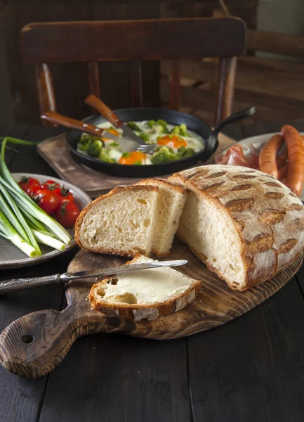 Breakfast is served on a black table: scrambled eggs, sausages, vegetables and homemade bread with butter. — Stock Photo, Image