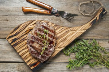 Bir kesme tahtası yatıyor ahşap bir masa üzerinde üst görünümü. Tahtada ızgara mermer et lisif var. Çatal bıçak takımı ve otların yakınında.