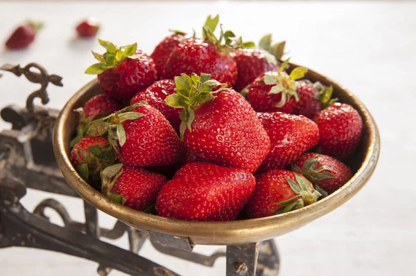 In a bowl of old scales that stand on a white table, lies a ripe strawberry.