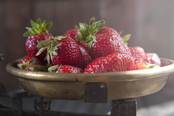 Sunlight falls on a copper bowl of cast-iron scales, in which a ripe strawberry lies.