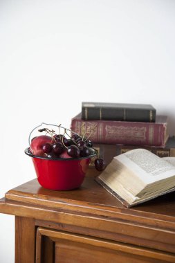 Çekmeceeski bir göğüs üzerinde eski kitaplar ve çilek ile kırmızı bir emaye kase vardır.