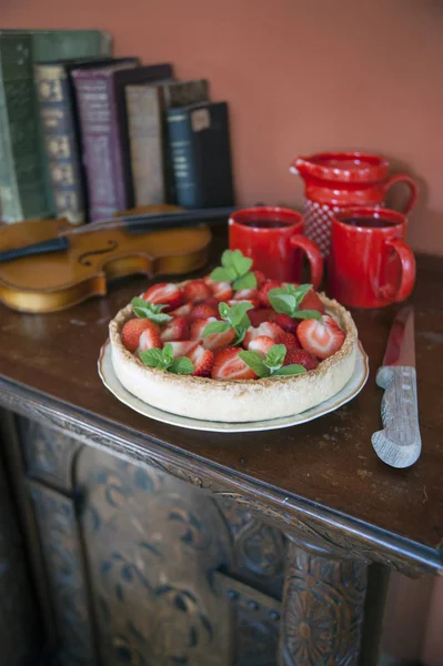 On an old carved dresser there is a plate with berry pie. Next to the knife is a red clay set. Behind lies a violin and old books.
