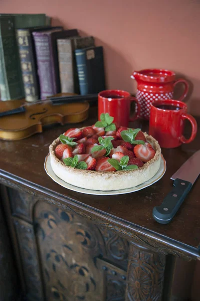 On an old carved dresser is a plate in which lies a berry pie. Next to the knife is a red clay set. Behind lies a violin and old books.