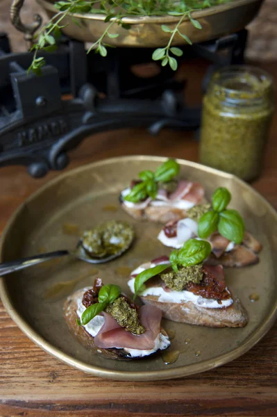 Sul vecchio tavolo c'è un piatto di rame con una bruschetta con crema di formaggio, pancetta, pomodori, pesto e basilico. Dietro ci sono scale antiche . — Foto Stock