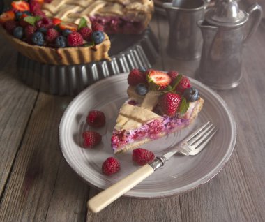 Güneş ışığı, ahşap bir masanın üzerindeki bir tabakta yer alan çileklerle süslenmiş bir dilim dut turtası aydınlatır. Pasta ve gümüş servisinin arkasında.