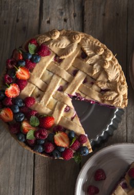 Taze ahududu ve çilek ile süslenmiş bir dut pasta standı üzerinde duruyor ahşap bir masa üzerinde üst görünümü. Tabağın yanında bir dilim kek ve çilek var..