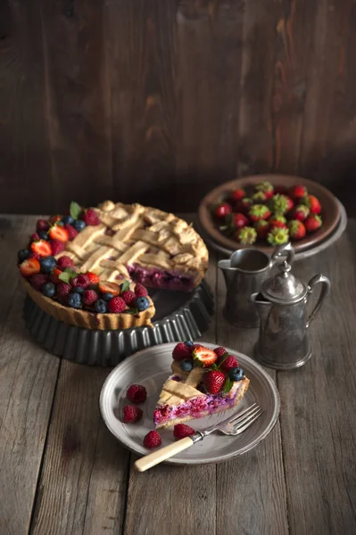 A slice of berry pie decorated with berries lies in a plate with a fork on a wooden table. Behind the cake, a silver service and plates with strawberries.