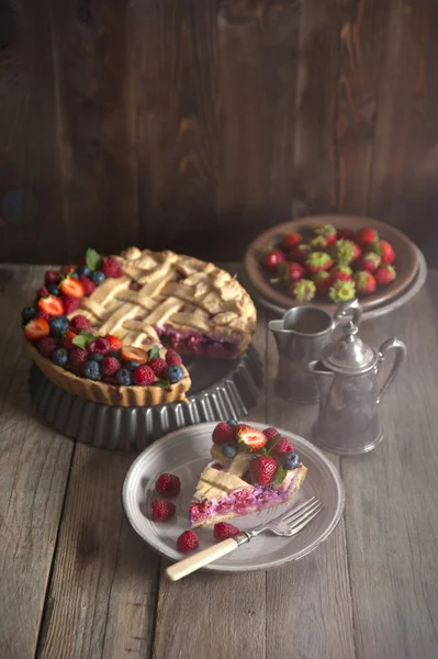 Sunlight illuminates a slice of berry pie decorated with berries, which lies in a plate with a fork on a wooden table. Behind the cake, a silver service and plates with strawberries.