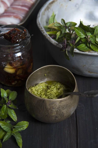 Su un tavolo di legno nero c'è un barattolo di rame con salsa di pesto e un cucchiaio. Vicino a un colabrodo con basilico, una scatola di pomodori asciugati al sole e una forma con rotoli di carne . — Foto Stock