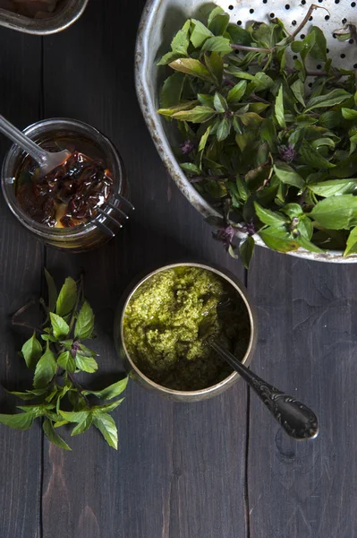 Draufsicht auf einen schwarzen Holztisch, auf dem ein Kupferglas mit Pesto-Sauce und einem Löffel steht. in der Nähe eines Siebes mit Basilikum und einem Glas getrockneter Tomaten. — Stockfoto