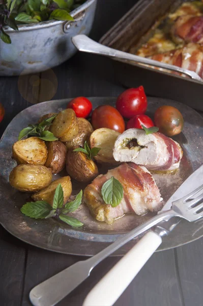 Sonnenlicht erhellt einen silbernen Teller mit Fleischbrötchen und mit Kräutern bestreutem Gemüse, der auf einem schwarzen Holztisch steht. hinter einer Backform mit Brötchen und einem Sieb mit grünem Basilikum. — Stockfoto