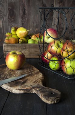 Metal bir sepet ve sandık siyah ahşap bir masa üzerinde elma ve armut vardır. Kesme tahtasının yanında.