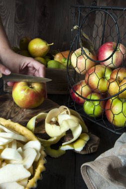 Pişirme için hazırlanan elma dolgulu bir pasta ile bir form masada. Yakınlarda metal bir sepet ve sandık ta elma ve armutbulunmaktadır. Bıçaklı kız kesme tahtasında meyve kesecek..