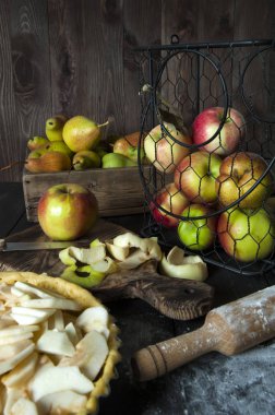Pişirme için hazırlanan elmalı turta ile bir tava masada. Oklavanın yanında ve kesme tahtasında elma soyma. Arkasında metal bir sepet ve sandık elma ve armut vardır.