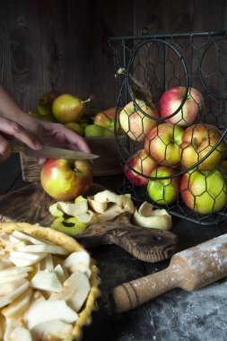 Pişirme için hazırlanan elmalı turta ile bir tava masada. Oklavanın yanında. Bıçaklı kız kesme tahtasında meyve kesecek. Bir sepet ve bir kutu arkasında elma ve armut vardır.