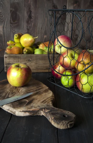 Metal bir sepet ve sandık siyah ahşap bir masa üzerinde elma ve armut vardır. Kesme tahtasının yanında.