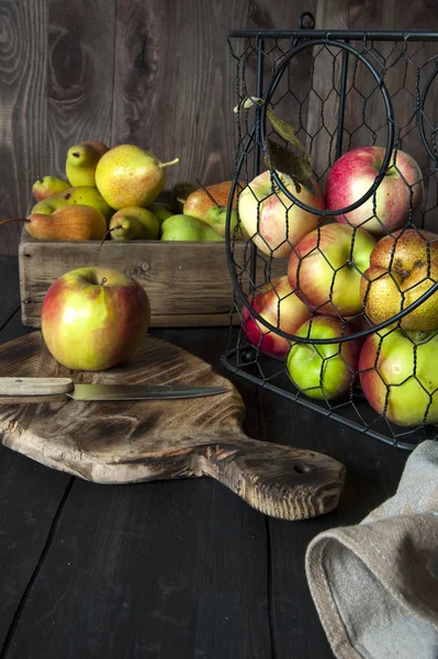 Ripe apples and pears are laid out in a basket and a box on a black wooden table. Nearby lies a cutting board with a knife and fruit. — Stock Photo, Image
