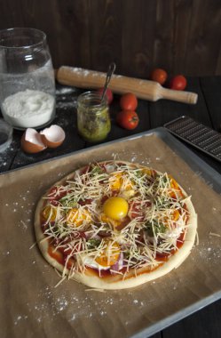 Un serpilir pişirme kağıdı ile kaplı bir fırın tepsisine siyah ahşap bir masa üzerinde, pişirme için hazırlanan bir pizza yatıyor. Oklava, domates, pesto ve un ile kavanoz arkasında.