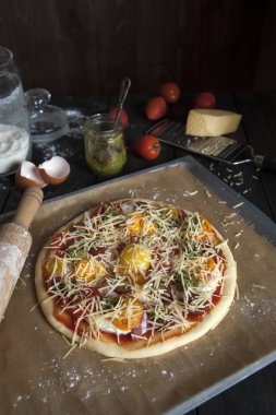 Fırın kağıdı ile kaplı bir fırın tepsisine siyah ahşap bir masa üzerinde pişirme için hazırlanan bir pizza yatıyor. Bir oklava, domates, pesto ve un ve parmesan ile bir rende ile kavanoz yakınında.