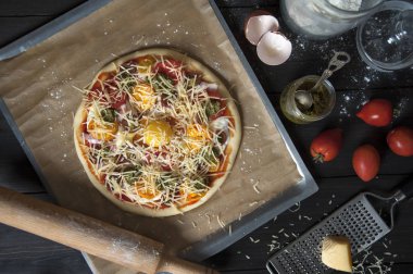 Pişirme kağıdı ile kaplı bir fırın tepsisine yatıyor pişirme için hazırlanan pizza üst görünümü. Bir oklava, yumurta kabuğu, domates ve pesto ve un ile kavanoz yakınında. Bir rende üzerinde Peynir.