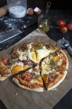 Dilimler halinde kesilmiş taze pişmiş pizza fırın kağıdı üzerinde siyah ahşap bir masa üzerinde yatıyor. Pizza bıçağının yanında. Domates arkasında, pesto ve un, yumurta kabuğu ve rende ile kavanoz.