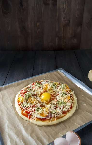 Fırın kağıdı ile kaplı bir fırın tepsisine siyah ahşap bir masa üzerinde pişirme için hazırlanan bir pizza yatıyor.