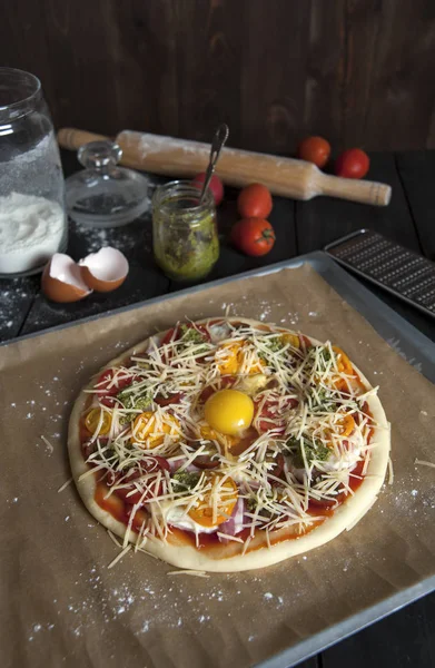 Un serpilir pişirme kağıdı ile kaplı bir fırın tepsisine siyah ahşap bir masa üzerinde, pişirme için hazırlanan bir pizza yatıyor. Bir oklava arkasında, domates, pesto ve un ve bir rende ile kavanoz.