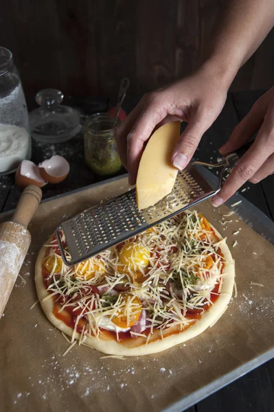 On a baking sheet covered with baking paper lies a pizza prepared for baking. Female hands on a grater tinder on cheese pizza. Near a rolling pin, eggshell and jars with pesto and flour.