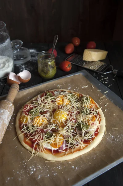Fırın kağıdı ile kaplı bir fırın tepsisine siyah ahşap bir masa üzerinde pişirme için hazırlanan bir pizza yatıyor. Bir oklava, domates, pesto ve un ve parmesan ile bir rende ile kavanoz yakınında.